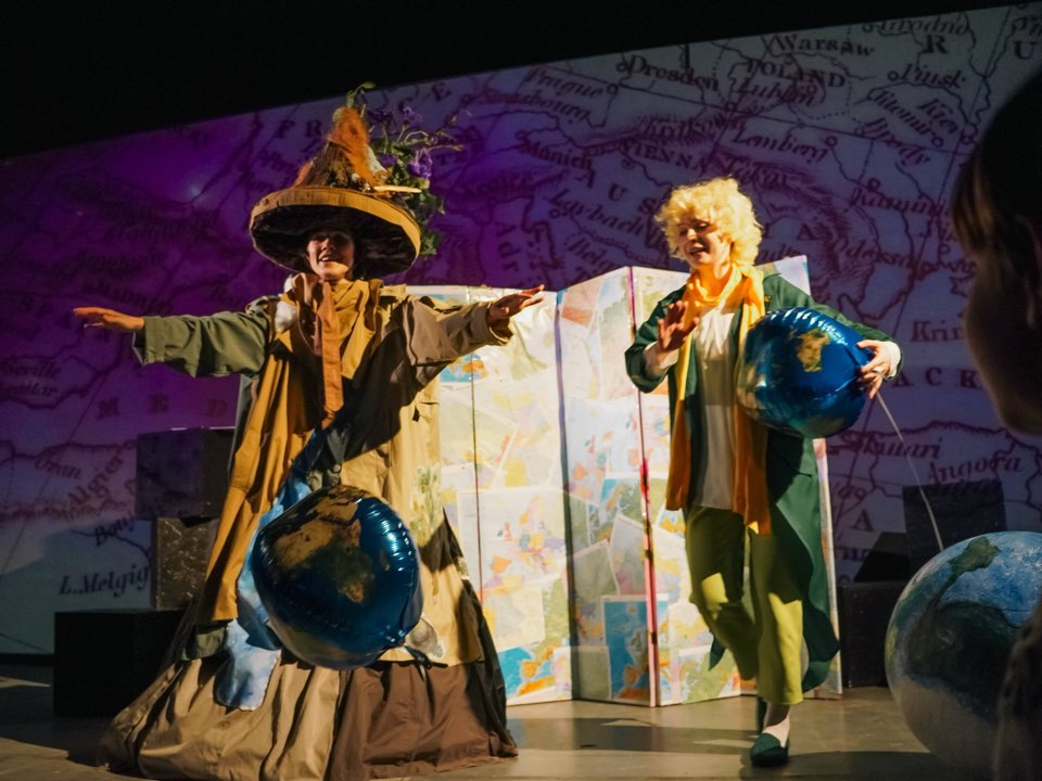 The Geographer holding a globe beside the Little Prince in front of a projected map