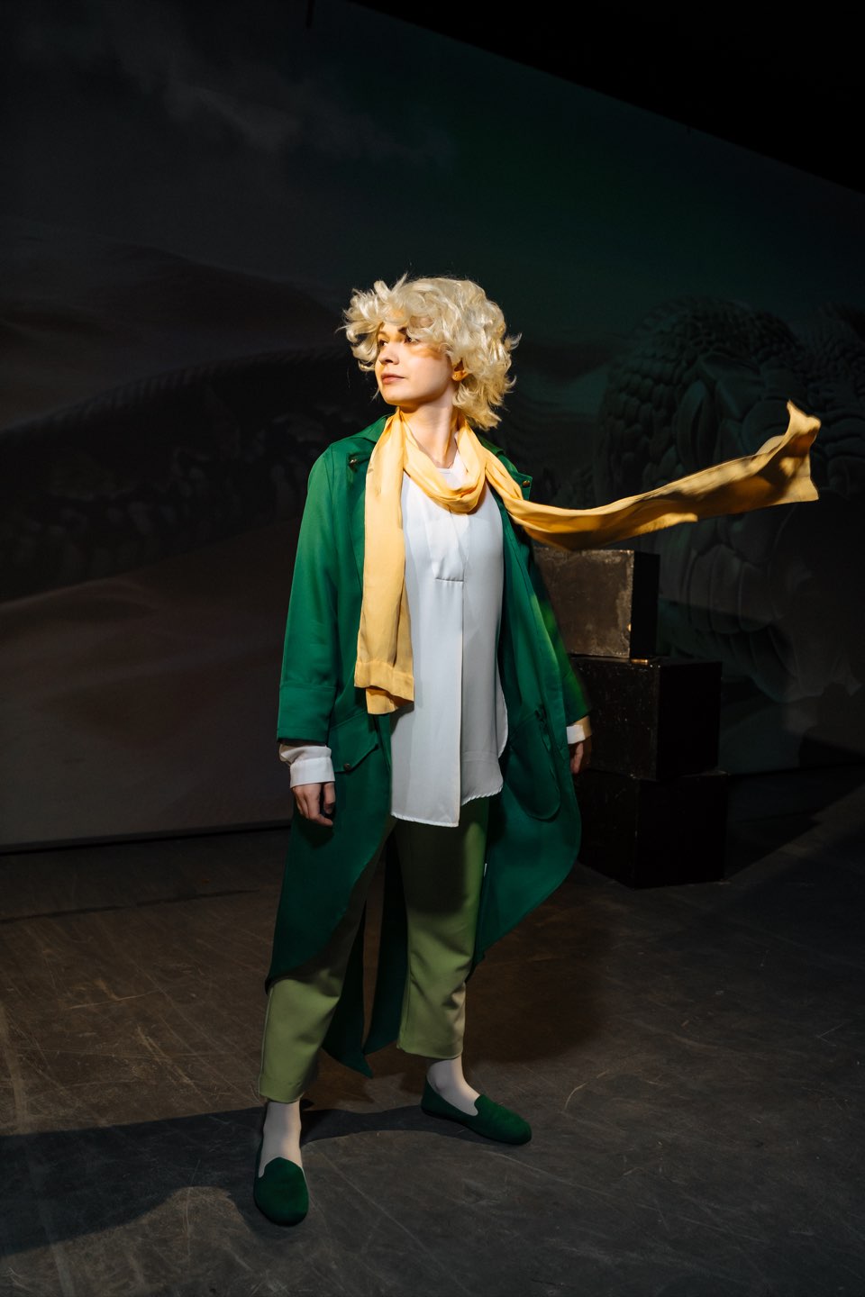 The Little Prince in a green coat with a yellow scarf blowing in the wind
